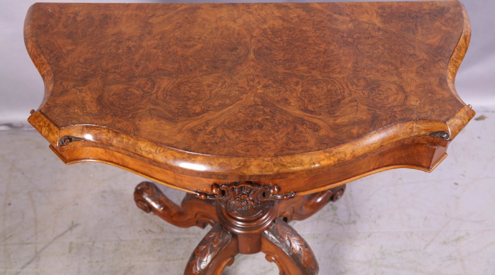 A Victorian burr walnut fold-over card table featuring a shaped top, a carved pedestal stem, and carved quadripartite cabriole legs on brass castors. View V