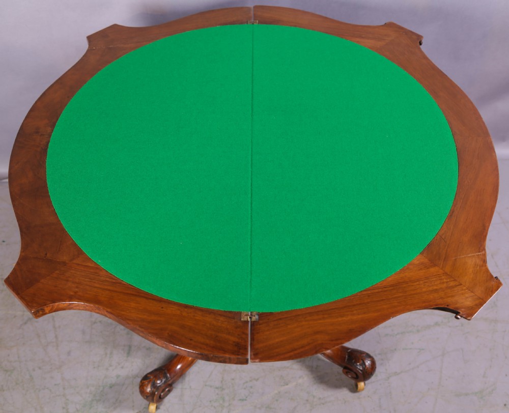 A Victorian burr walnut fold-over card table featuring a shaped top, a carved pedestal stem, and carved quadripartite cabriole legs on brass castors. View VI