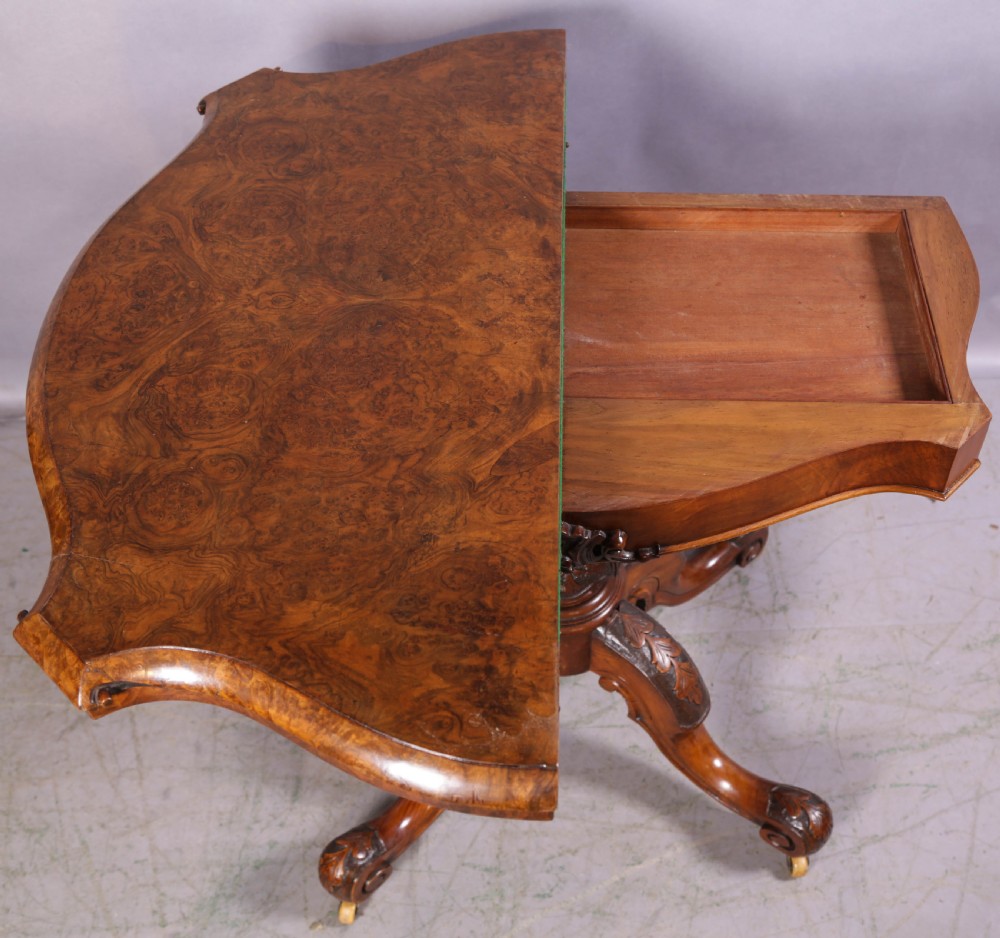 A Victorian burr walnut fold-over card table featuring a shaped top, a carved pedestal stem, and carved quadripartite cabriole legs on brass castors. View VIII