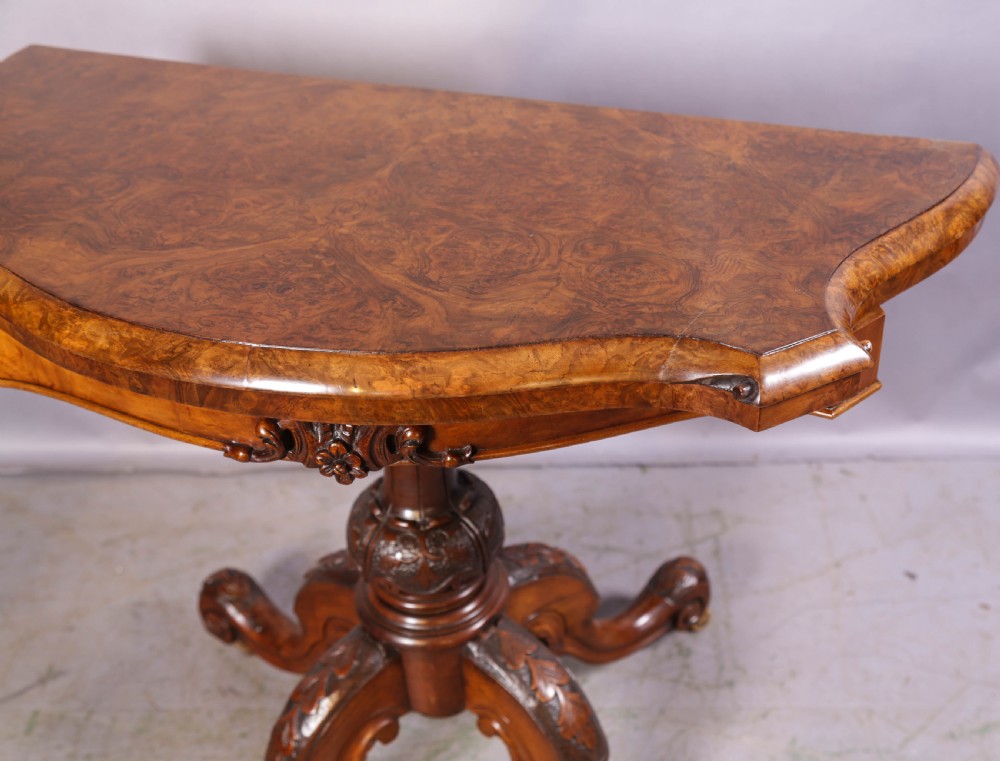 A Victorian burr walnut fold-over card table featuring a shaped top, a carved pedestal stem, and carved quadripartite cabriole legs on brass castors. View IX