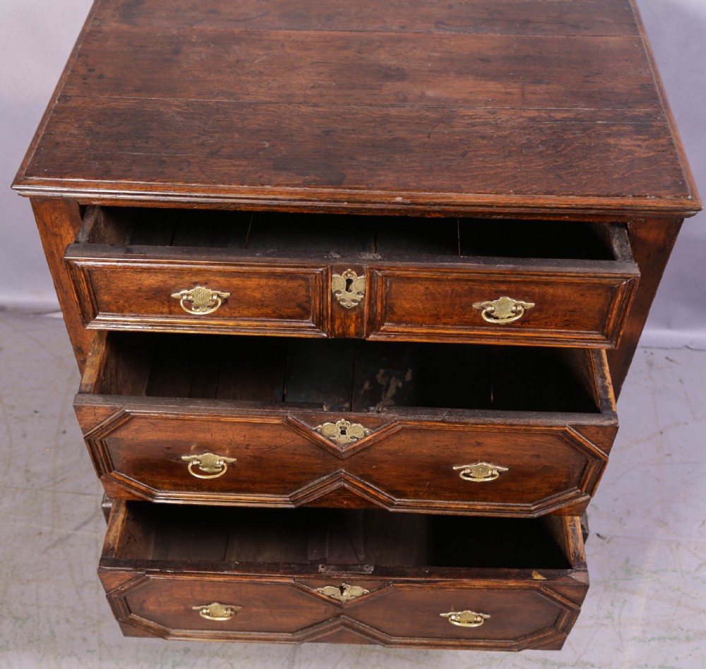 An early 18th-century dark oak chest of drawers featuring geometric moulding on the drawer fronts and brass drop handles. View VI