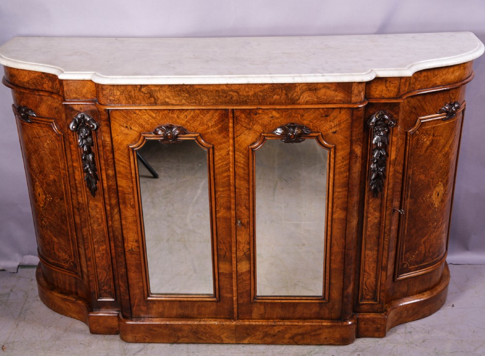 A serpentine-fronted Victorian burr walnut credenza featuring a white marble top, mirrored doors, and carved wooden mounts.