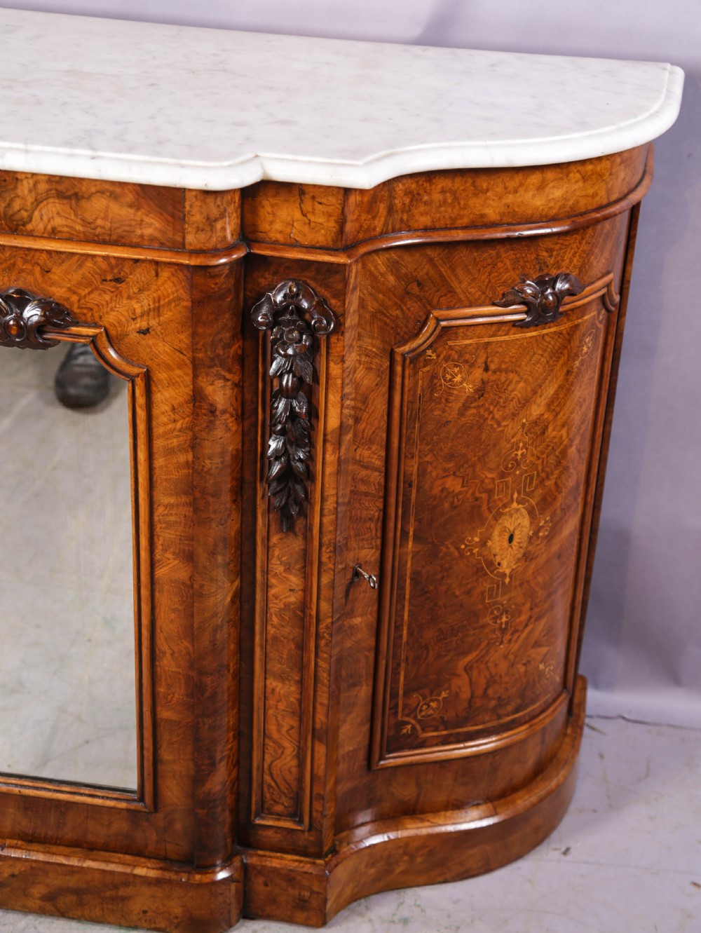 A serpentine-fronted Victorian burr walnut credenza featuring a white marble top, mirrored doors, and carved wooden mounts. View IV