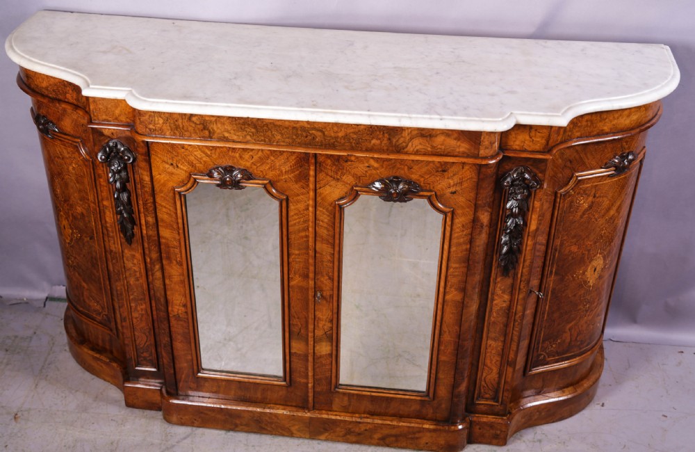 A serpentine-fronted Victorian burr walnut credenza featuring a white marble top, mirrored doors, and carved wooden mounts. View VI