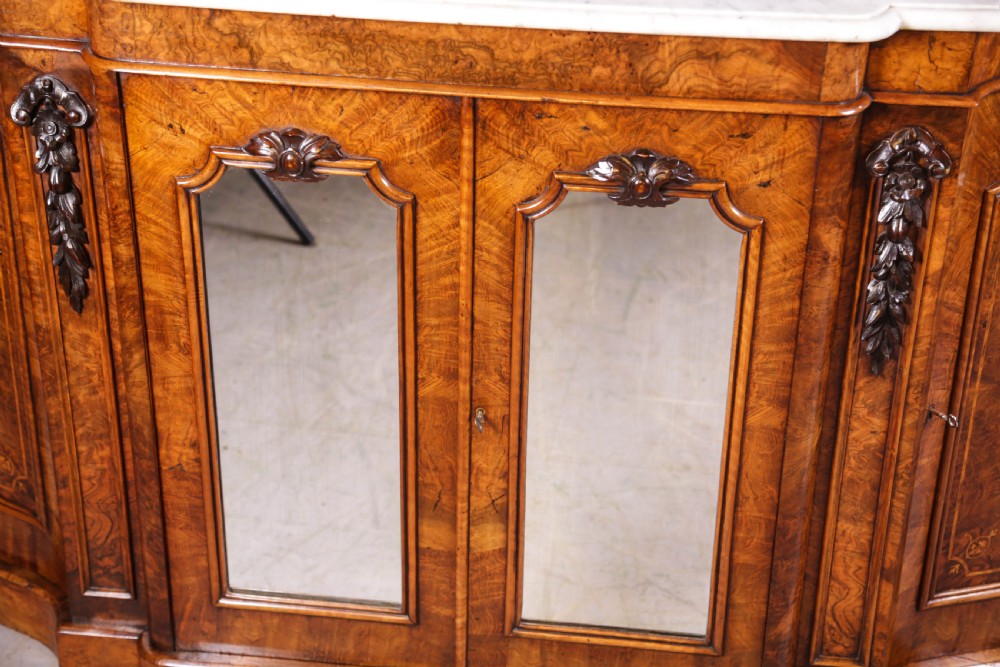 A serpentine-fronted Victorian burr walnut credenza featuring a white marble top, mirrored doors, and carved wooden mounts. View VII