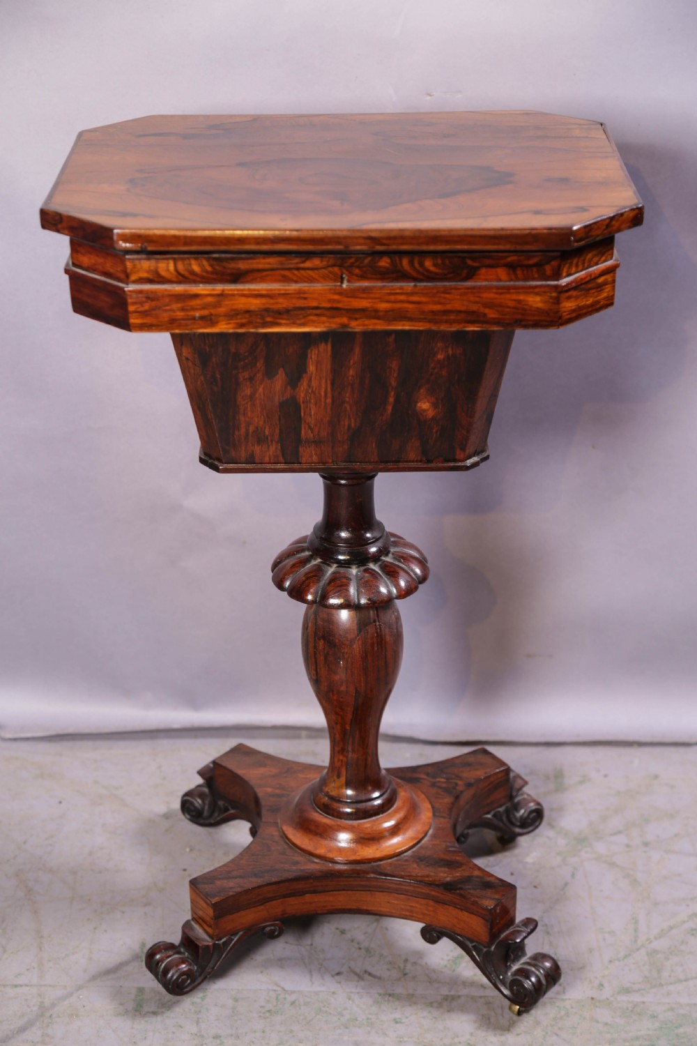 An octagonal Victorian rosewood sewing stand with a tapered compartment, a carved pedestal base, and scrolling feet on castors.