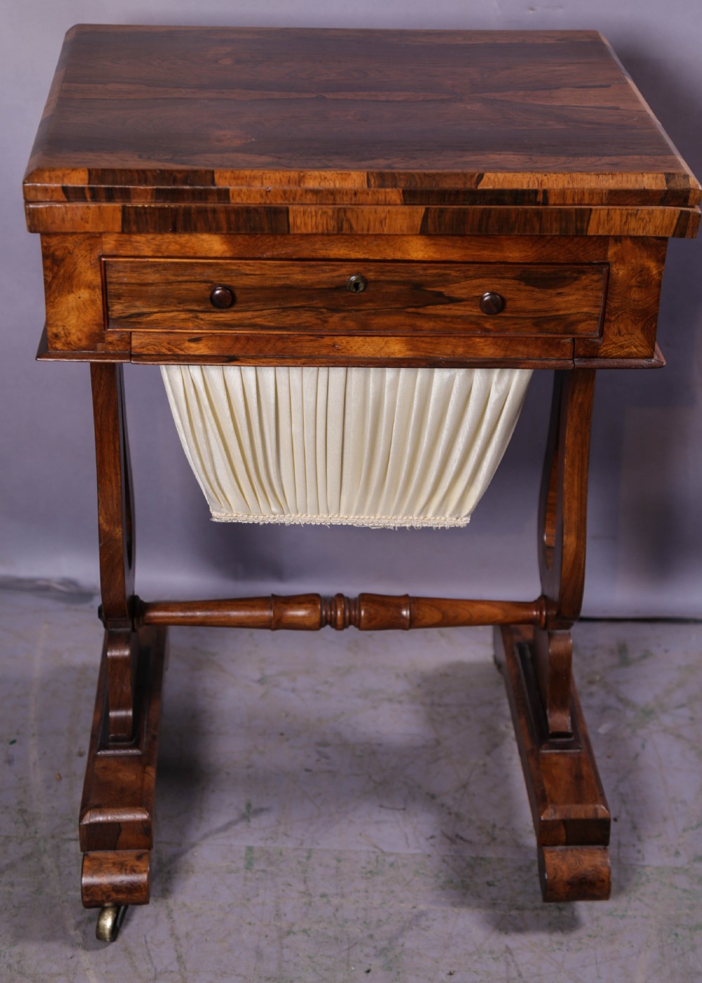 A Victorian rosewood sewing and games table featuring a fold-over top, a single frieze drawer, and a suspended pleated fabric work bag supported by a lyre-form pedestal base with brass castors.