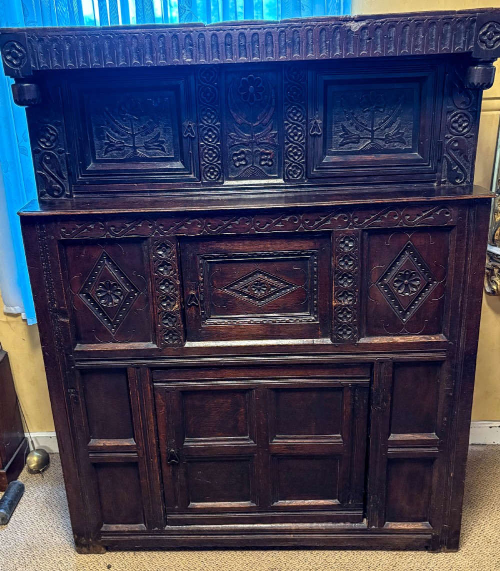 A front view of a dark brown antique oak livery cupboard featuring elaborate hand-carved geometric panels, friezes, and cupboard doors.