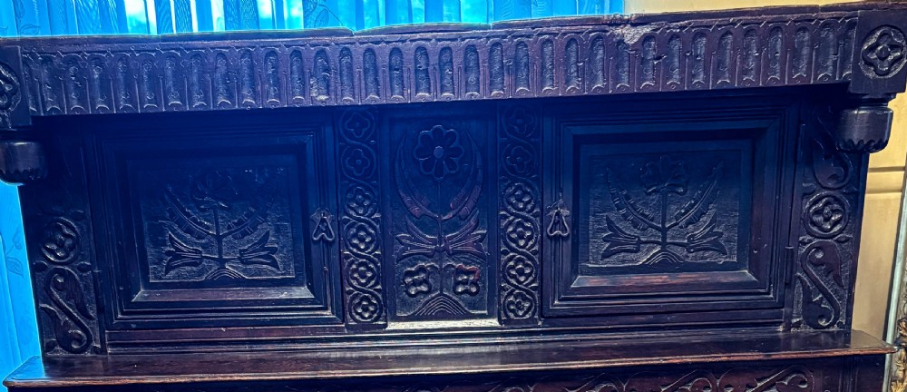 A front view of a dark brown antique oak livery cupboard featuring elaborate hand-carved geometric panels, friezes, and cupboard doors. View II