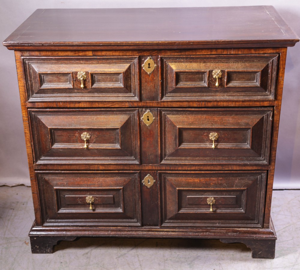 A dark brown George I oak chest of drawers featuring geometric panelled drawer fronts and brass drop handles.