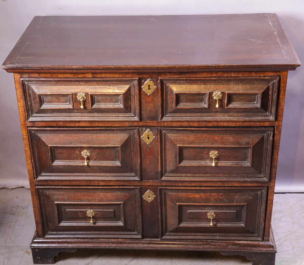 A dark brown George I oak chest of drawers featuring geometric panelled drawer fronts and brass drop handles. View II
