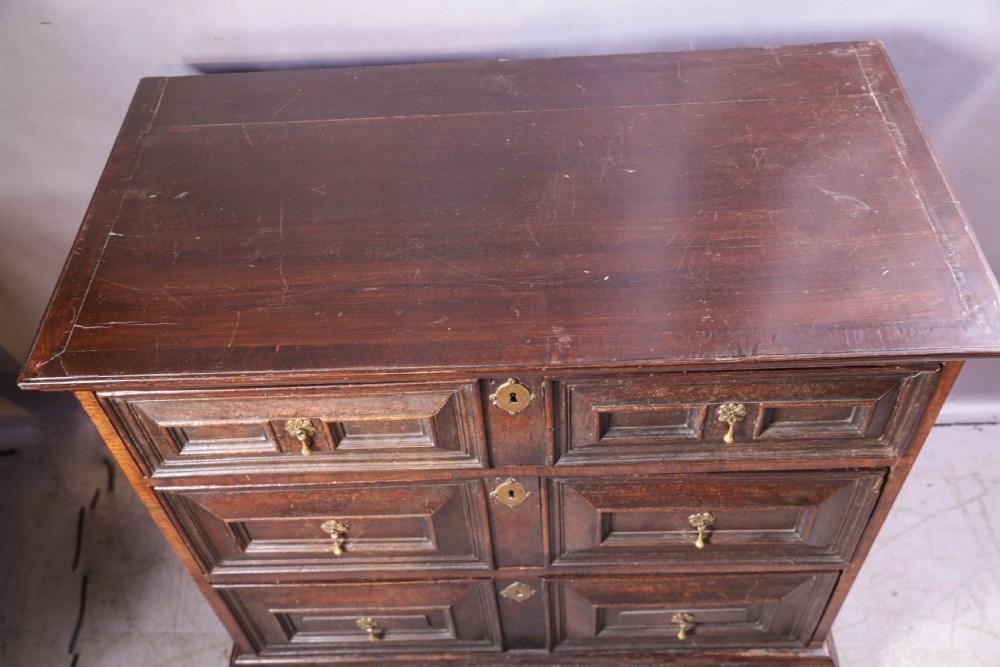 A dark brown George I oak chest of drawers featuring geometric panelled drawer fronts and brass drop handles. View IV