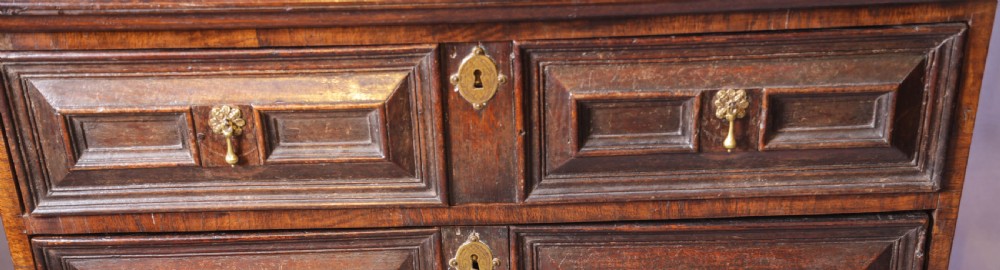 A dark brown George I oak chest of drawers featuring geometric panelled drawer fronts and brass drop handles. View V