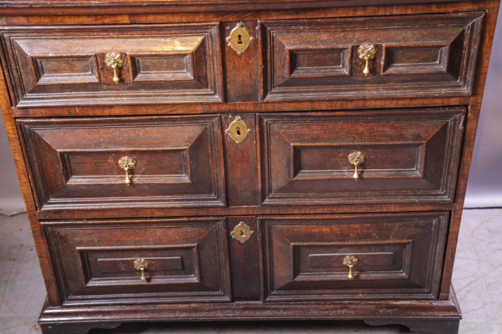 A dark brown George I oak chest of drawers featuring geometric panelled drawer fronts and brass drop handles. View VI