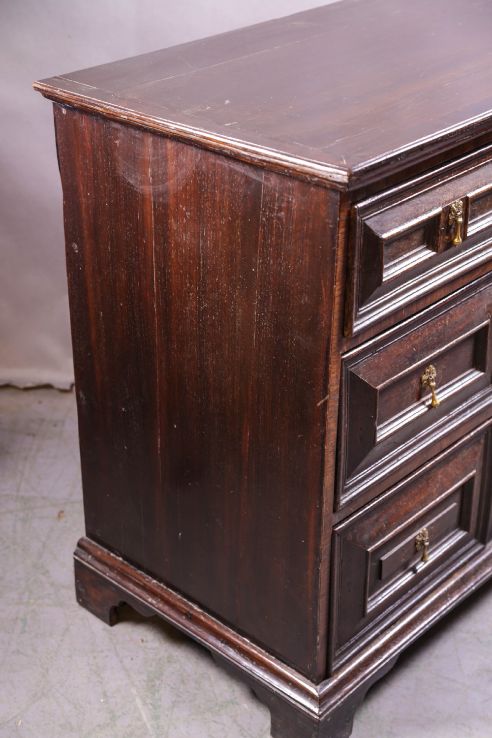 A dark brown George I oak chest of drawers featuring geometric panelled drawer fronts and brass drop handles. View VII