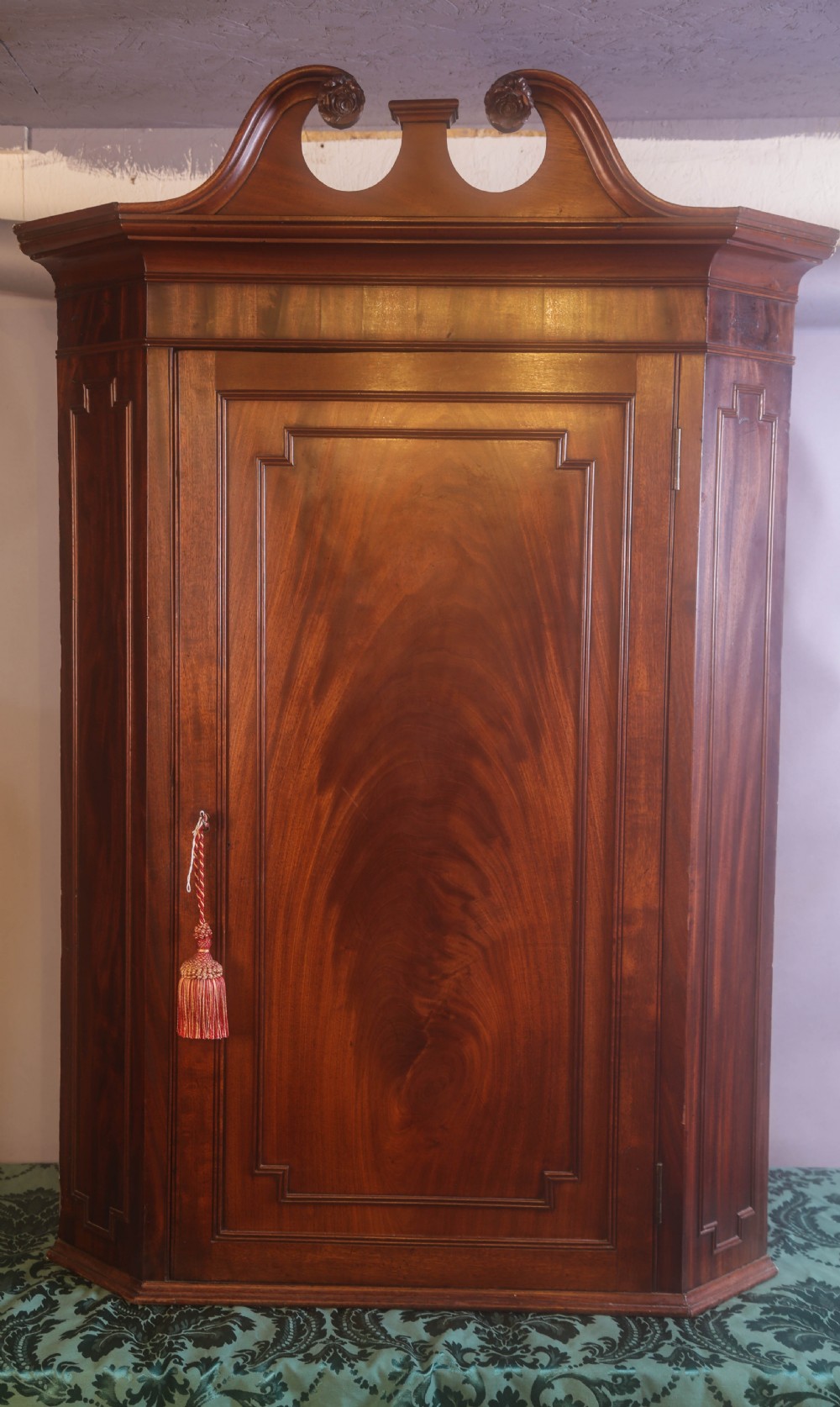 An antique Georgian flame mahogany corner cupboard featuring a swan-neck pediment with carved rosettes and a single panelled door.