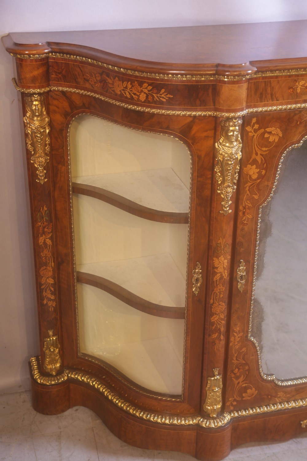 A Victorian burr walnut credenza featuring floral marquetry inlay, gilt-metal mounts, a central mirrored door, and curved glass side display compartments. View V