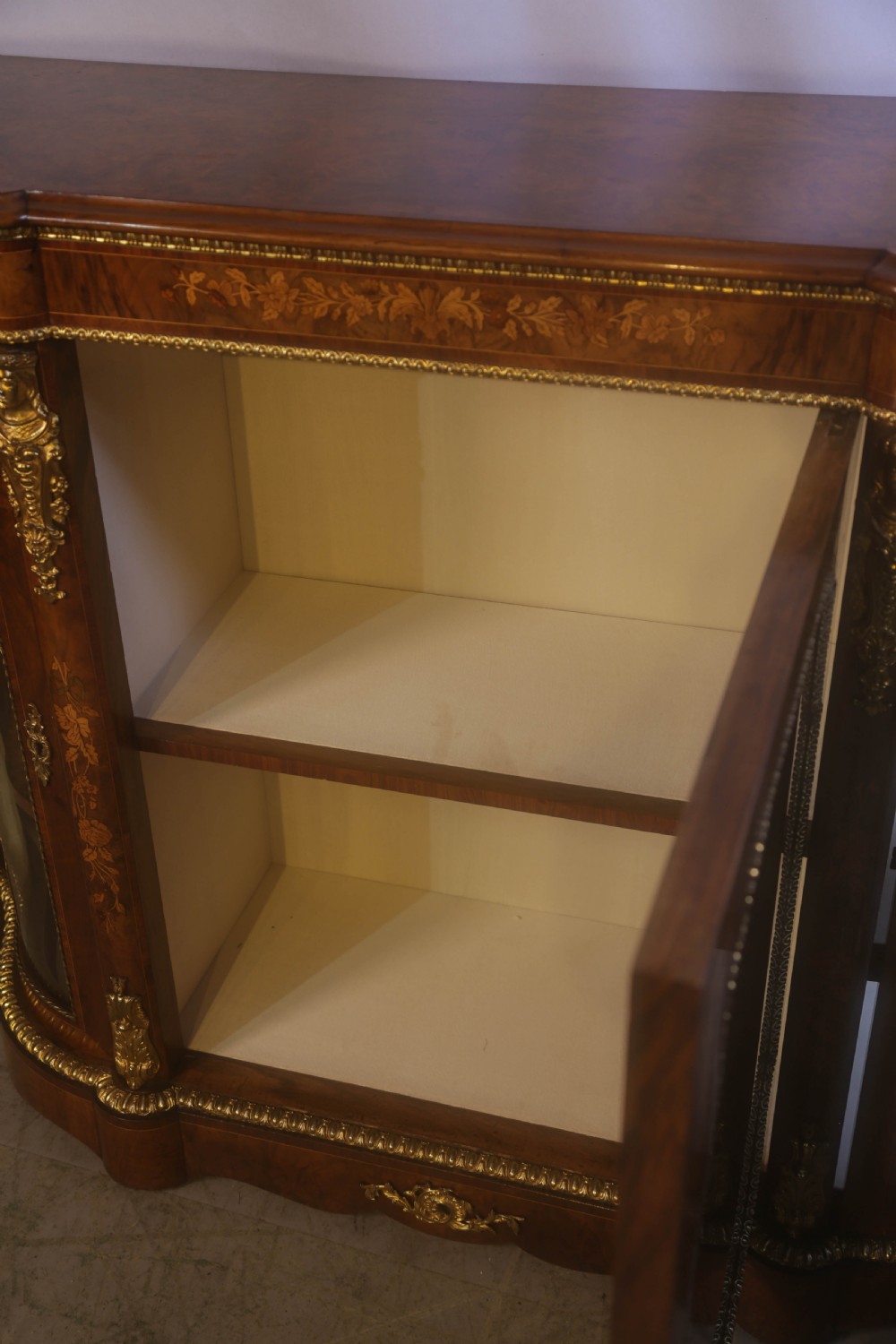A Victorian burr walnut credenza featuring floral marquetry inlay, gilt-metal mounts, a central mirrored door, and curved glass side display compartments. View IX