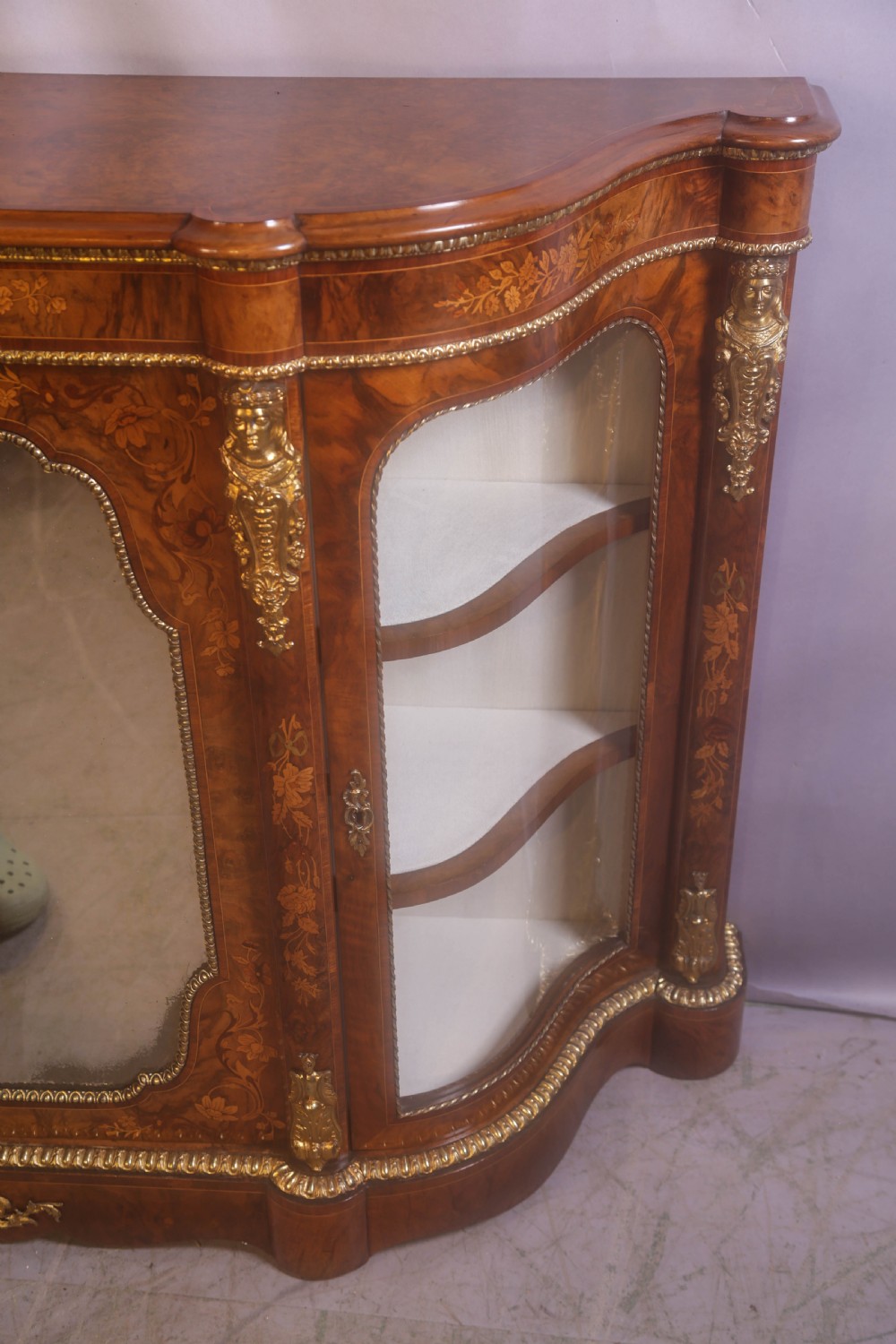 A Victorian burr walnut credenza featuring floral marquetry inlay, gilt-metal mounts, a central mirrored door, and curved glass side display compartments. View X