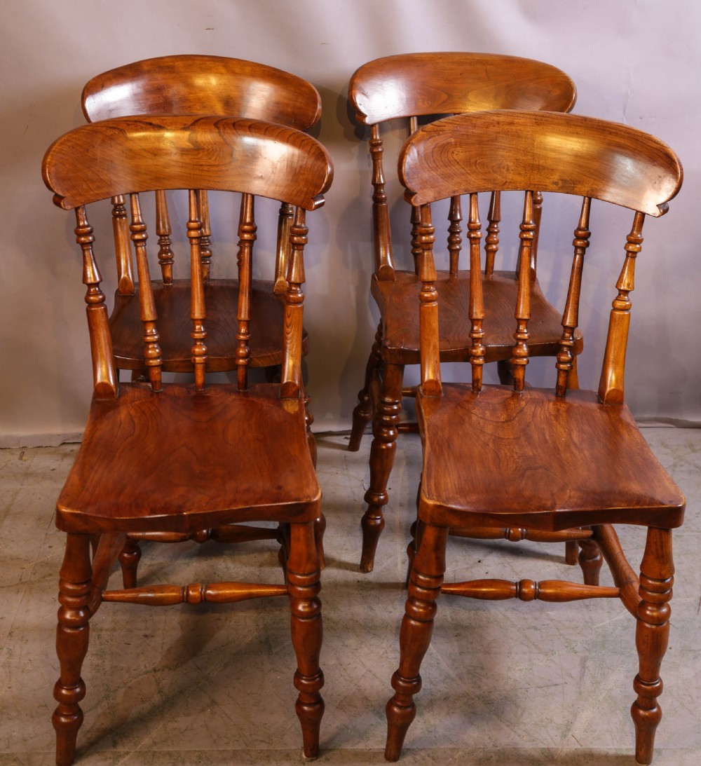 A set of four antique wooden kitchen chairs featuring rounded crest rails, turned back spindles, shaped solid seats, and turned legs.