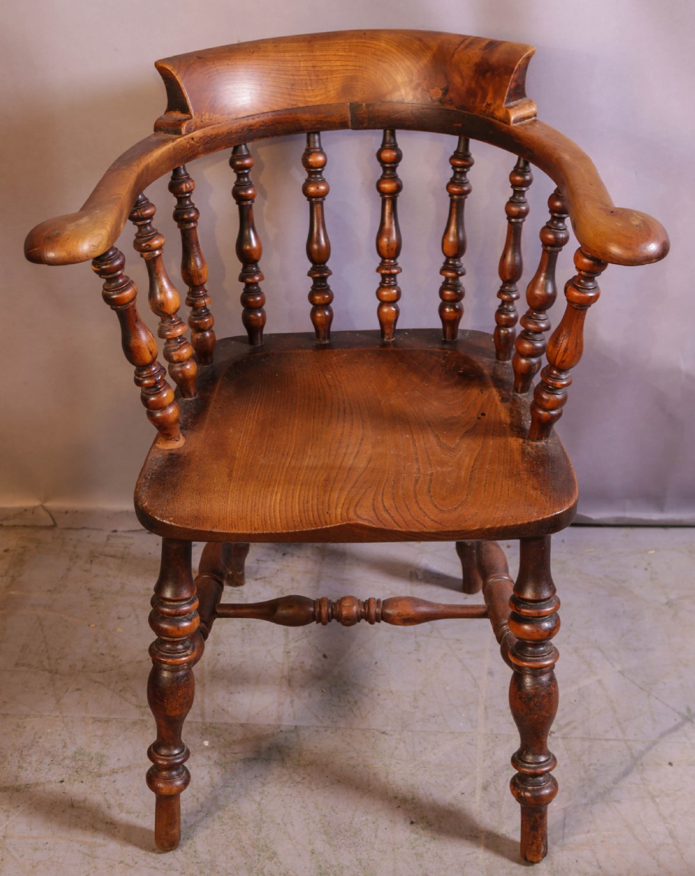 A Victorian captains chair made of polished yew wood featuring a curved crest rail, turned spindles, turned legs, and a solid shaped elm seat.