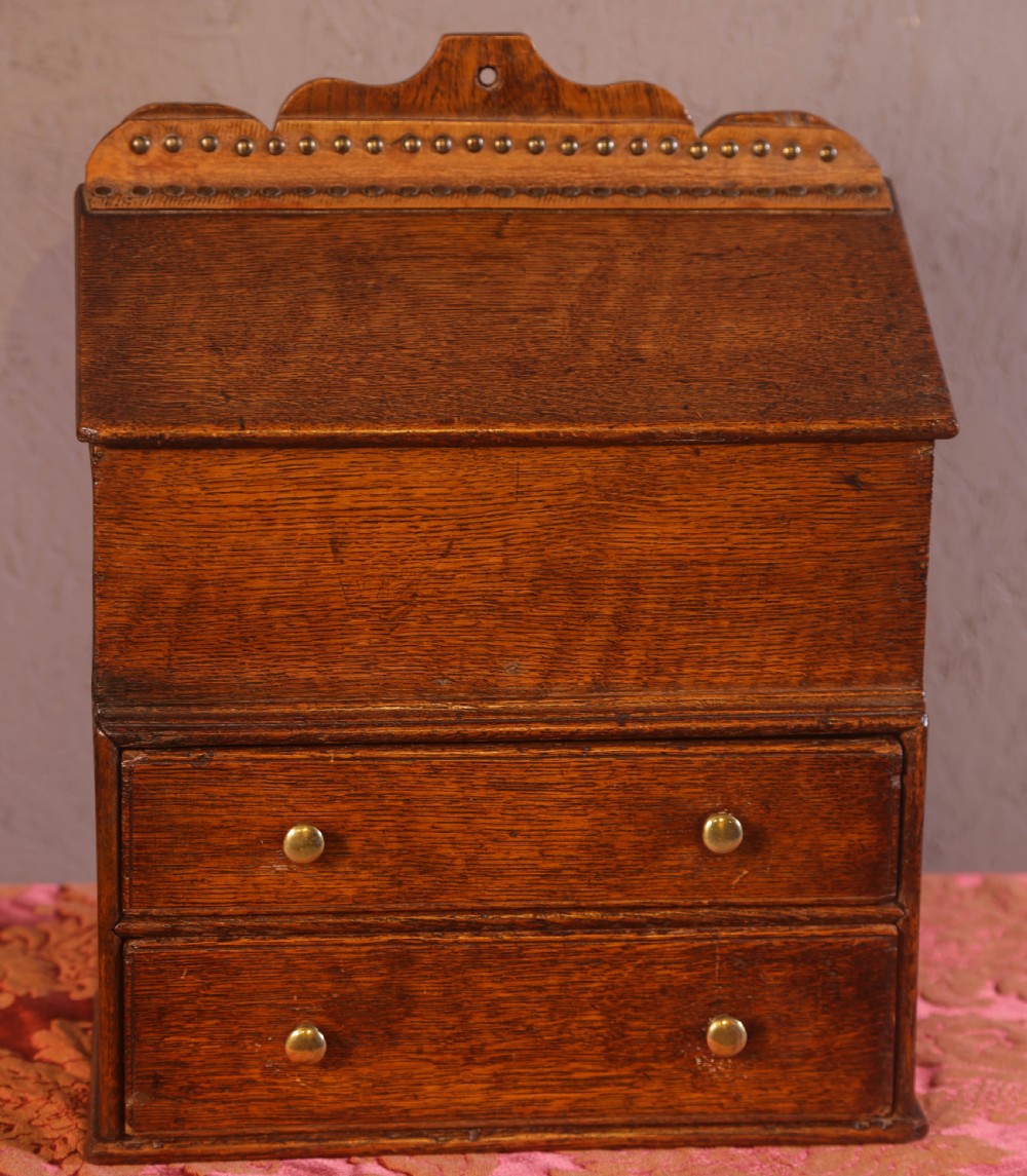 An antique warm brown oak candle box featuring a sloped lid, brass stud details on the shaped crest, and two drawers with brass knobs.