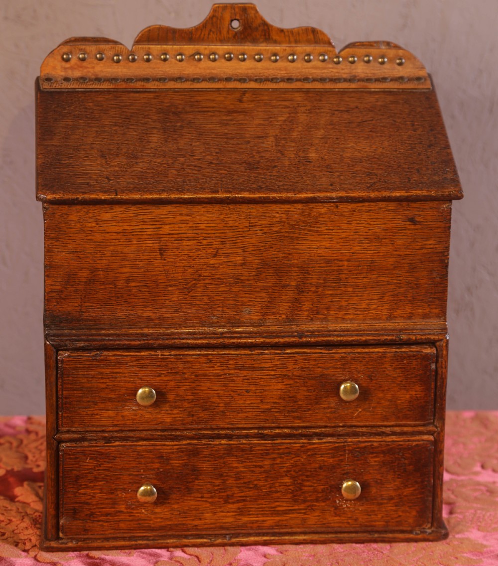 An antique warm brown oak candle box featuring a sloped lid, brass stud details on the shaped crest, and two drawers with brass knobs. View II
