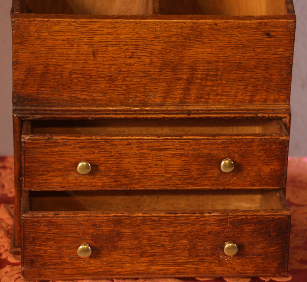 An antique warm brown oak candle box featuring a sloped lid, brass stud details on the shaped crest, and two drawers with brass knobs. View IV