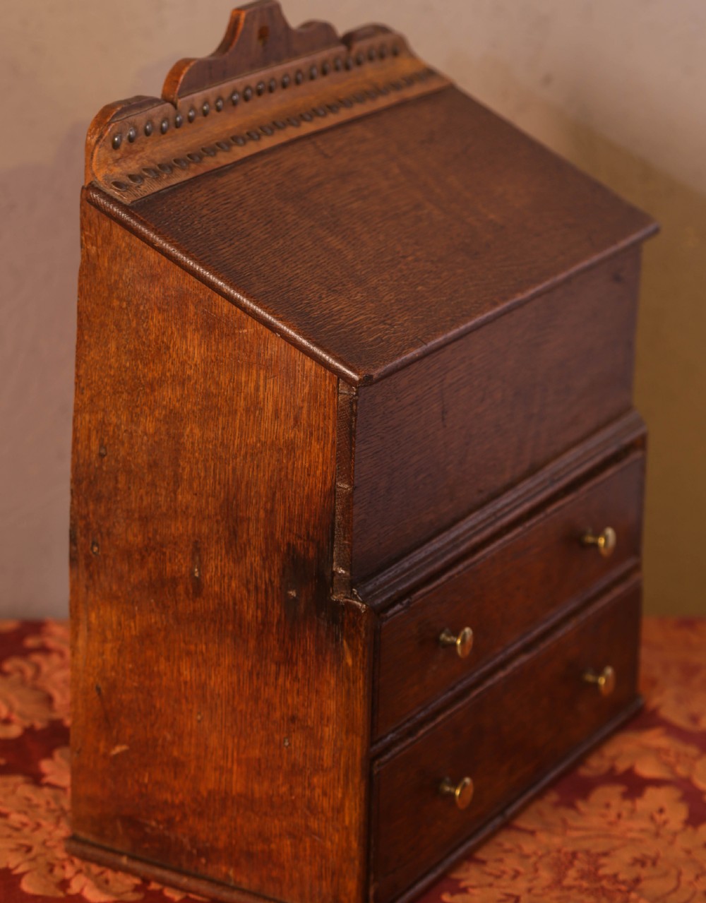An antique warm brown oak candle box featuring a sloped lid, brass stud details on the shaped crest, and two drawers with brass knobs. View VI