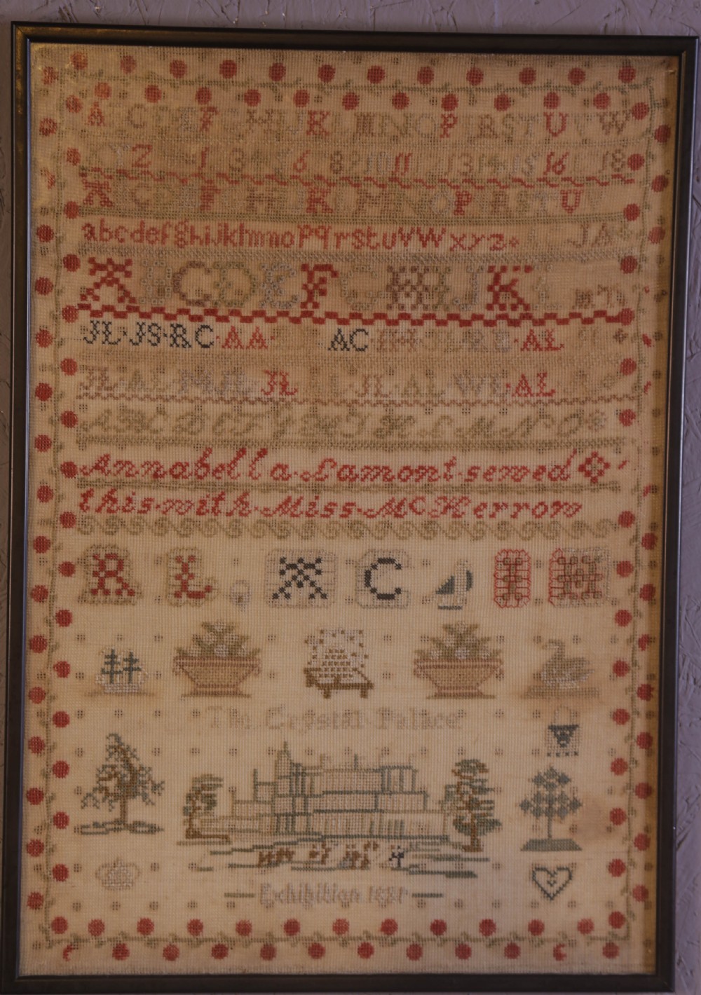 Framed 1851 Victorian needlework sampler featuring alphabets, numbers, and an embroidered depiction of the Crystal Palace on a linen ground.