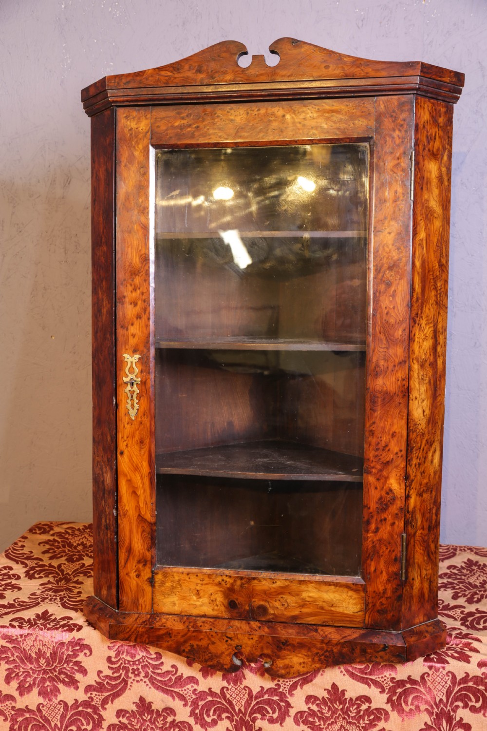 An antique corner cabinet crafted from figured yew wood featuring a single glazed door, decorative scroll pediment, and brass escutcheon.