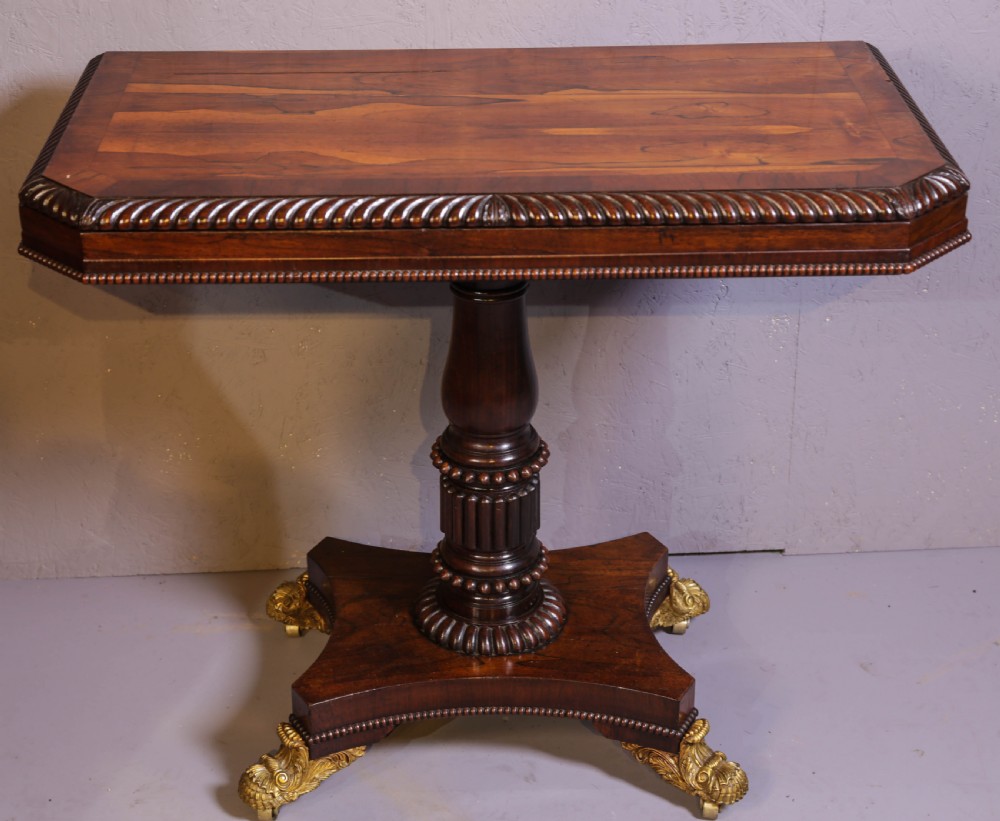 An antique Regency rosewood games table featuring a fold-over top with gadrooned trim, a turned pedestal support, and a quadform base with brass lion paw feet.