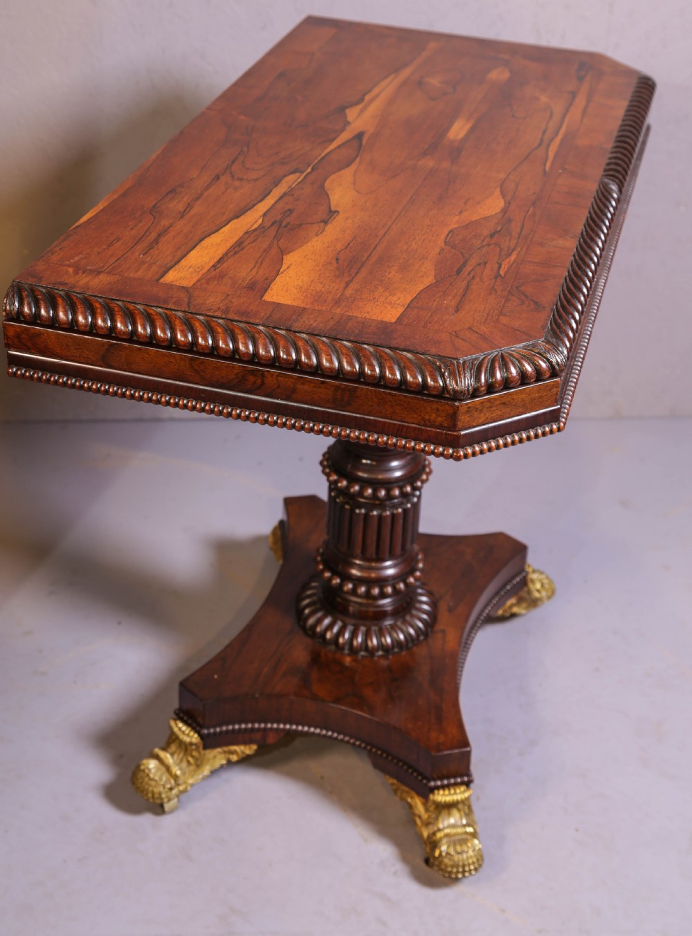 An antique Regency rosewood games table featuring a fold-over top with gadrooned trim, a turned pedestal support, and a quadform base with brass lion paw feet. View II