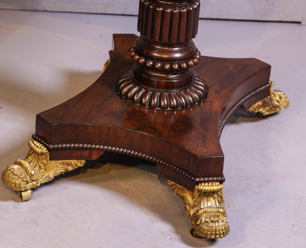 An antique Regency rosewood games table featuring a fold-over top with gadrooned trim, a turned pedestal support, and a quadform base with brass lion paw feet. View IV
