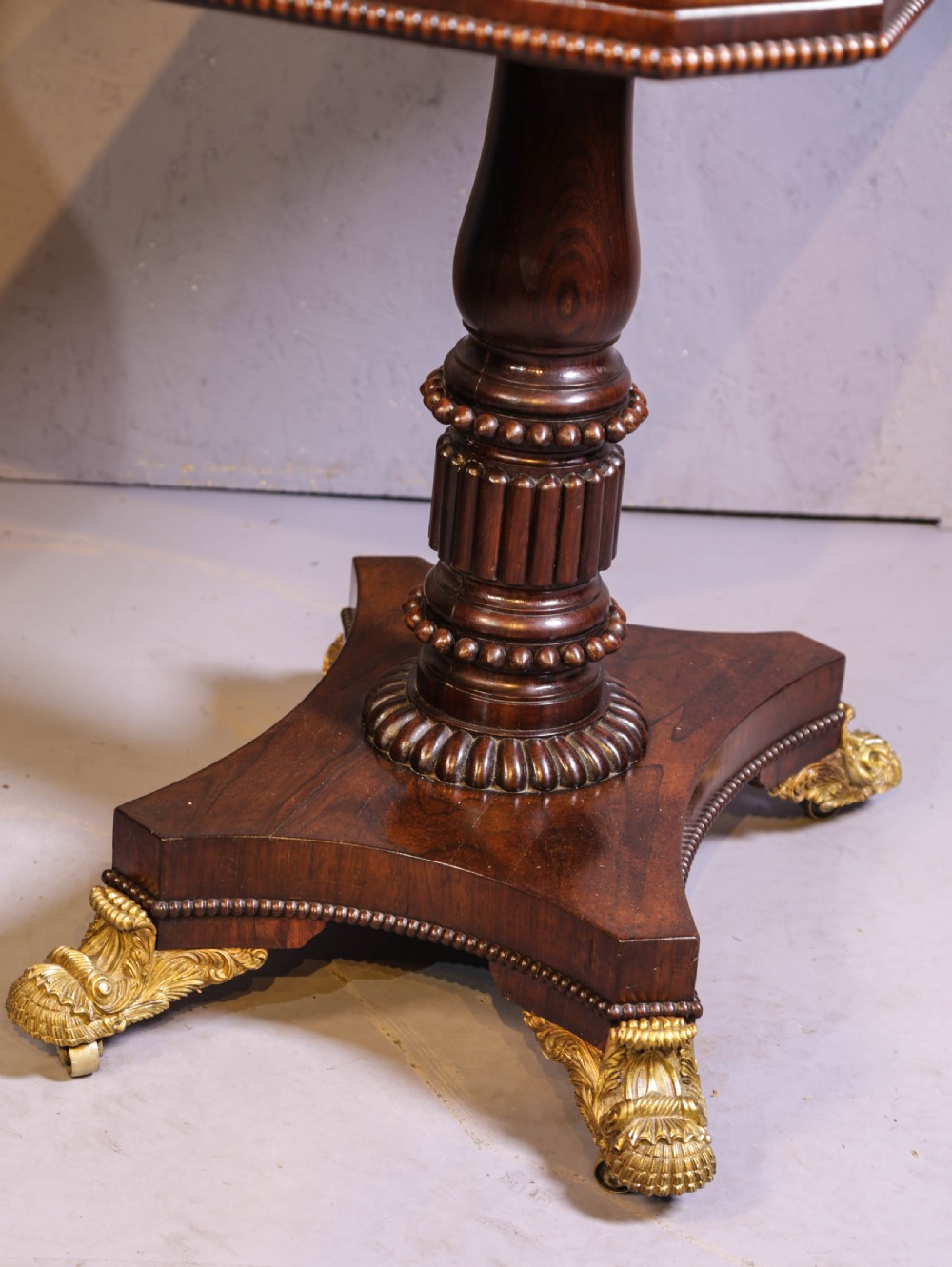 An antique Regency rosewood games table featuring a fold-over top with gadrooned trim, a turned pedestal support, and a quadform base with brass lion paw feet. View VI