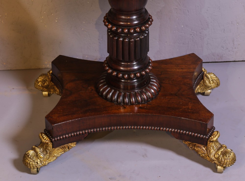 An antique Regency rosewood games table featuring a fold-over top with gadrooned trim, a turned pedestal support, and a quadform base with brass lion paw feet. View VII