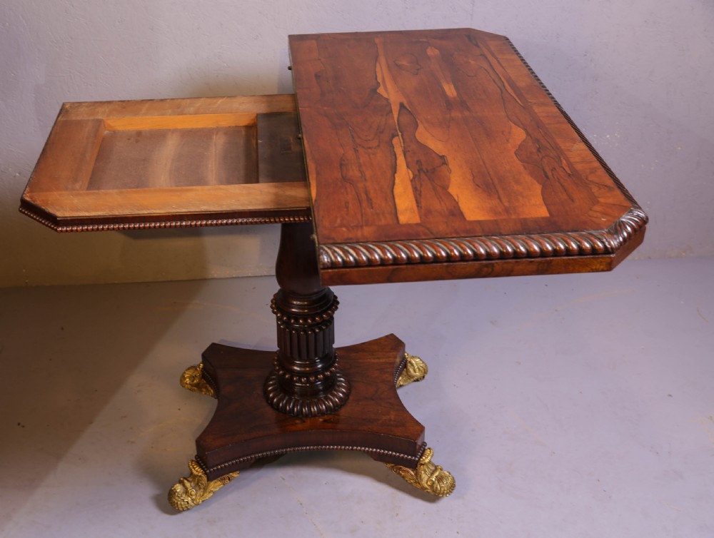 An antique Regency rosewood games table featuring a fold-over top with gadrooned trim, a turned pedestal support, and a quadform base with brass lion paw feet. View VIII