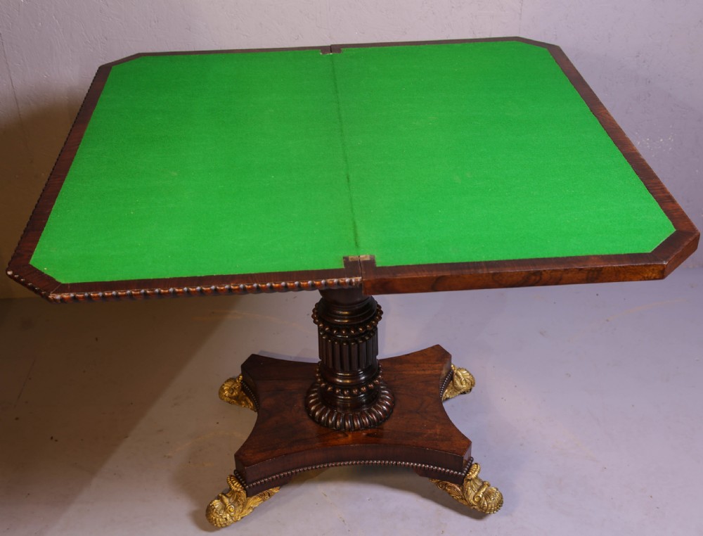 An antique Regency rosewood games table featuring a fold-over top with gadrooned trim, a turned pedestal support, and a quadform base with brass lion paw feet. View IX