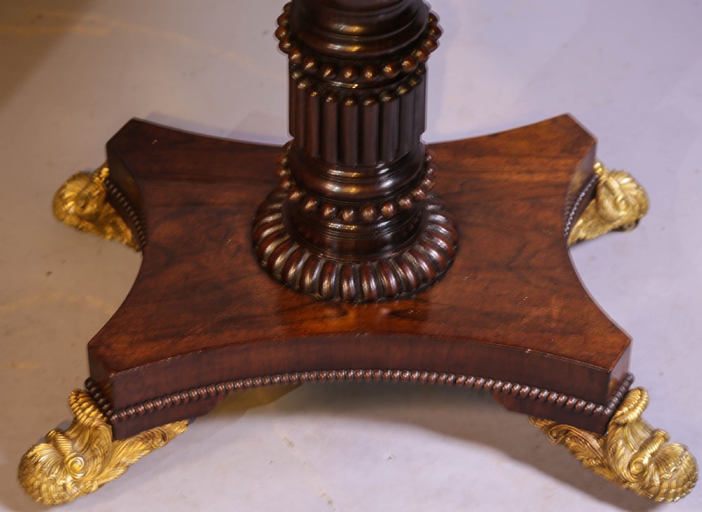 An antique Regency rosewood games table featuring a fold-over top with gadrooned trim, a turned pedestal support, and a quadform base with brass lion paw feet. View X