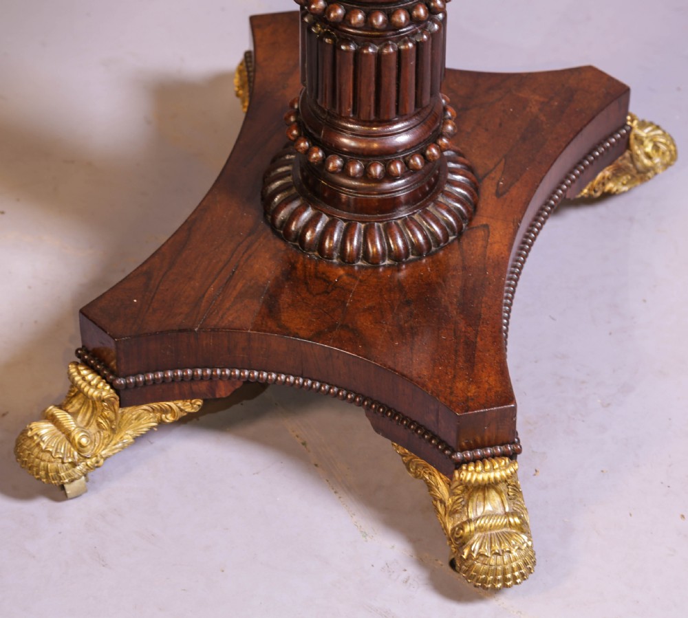 An antique Regency rosewood games table featuring a fold-over top with gadrooned trim, a turned pedestal support, and a quadform base with brass lion paw feet. View XI