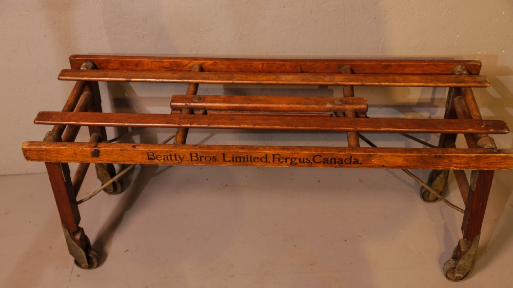 A slatted warm brown wooden luggage stand featuring metal castors and stamped maker details along the front rail.