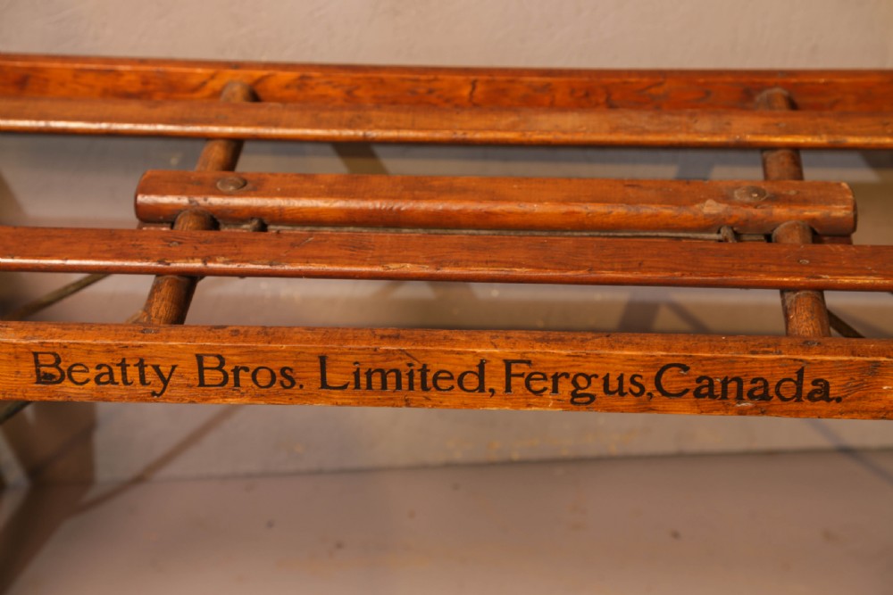 A slatted warm brown wooden luggage stand featuring metal castors and stamped maker details along the front rail. View II