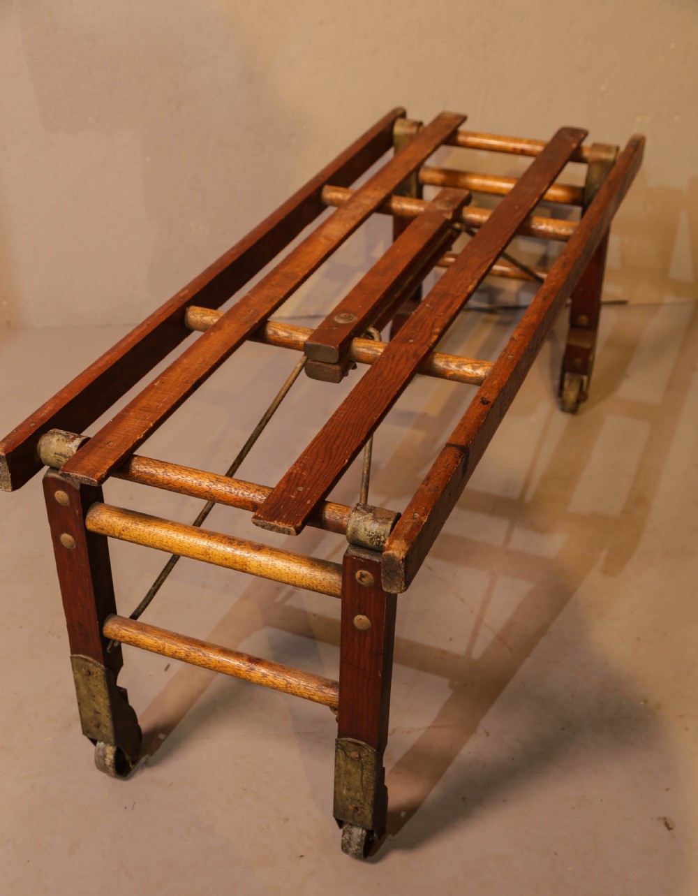 A slatted warm brown wooden luggage stand featuring metal castors and stamped maker details along the front rail. View III