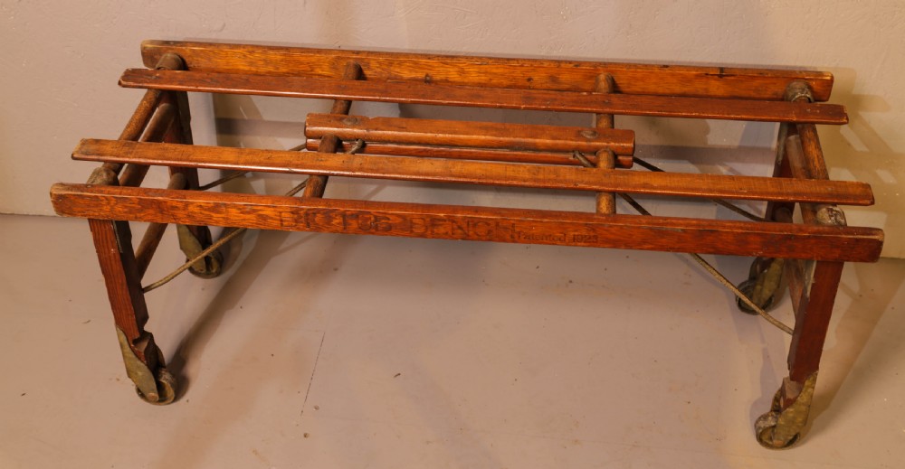 A slatted warm brown wooden luggage stand featuring metal castors and stamped maker details along the front rail. View IV