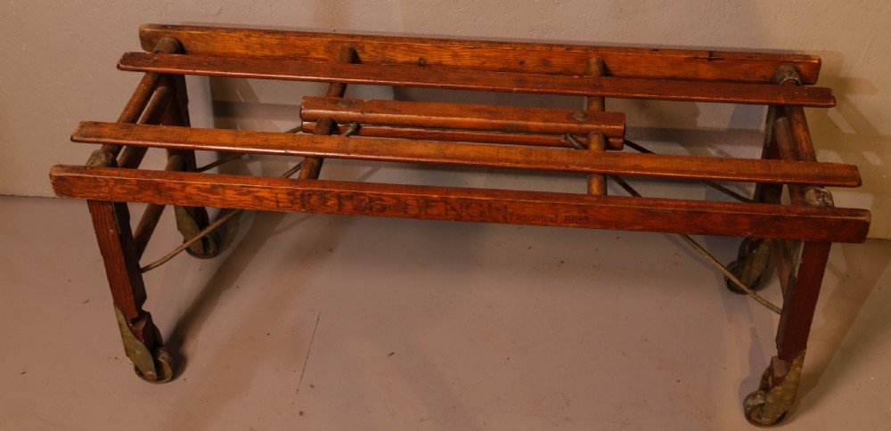A slatted warm brown wooden luggage stand featuring metal castors and stamped maker details along the front rail. View V