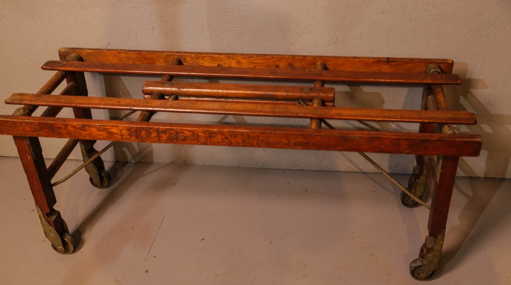 A slatted warm brown wooden luggage stand featuring metal castors and stamped maker details along the front rail. View VI