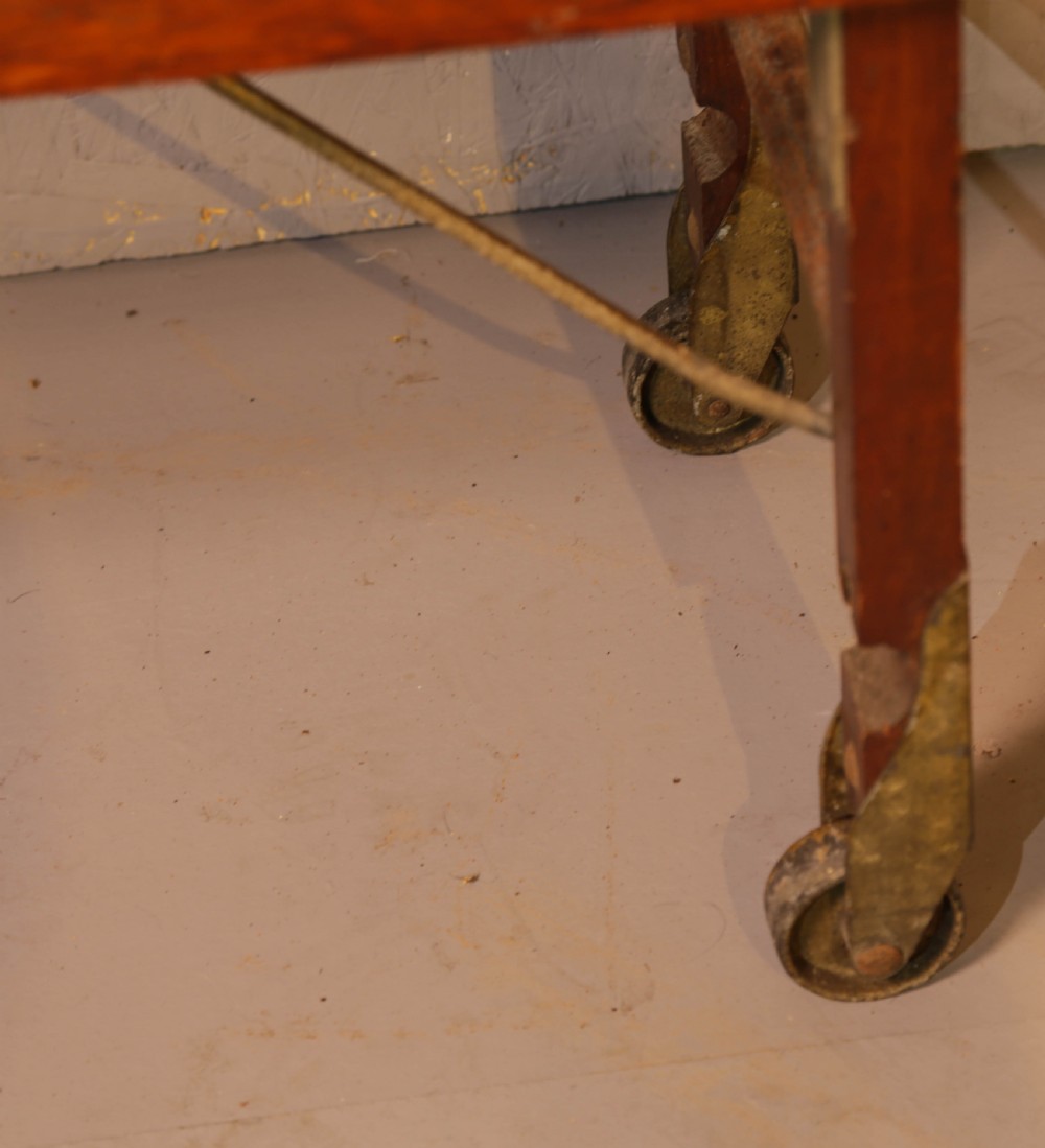 A slatted warm brown wooden luggage stand featuring metal castors and stamped maker details along the front rail. View VII