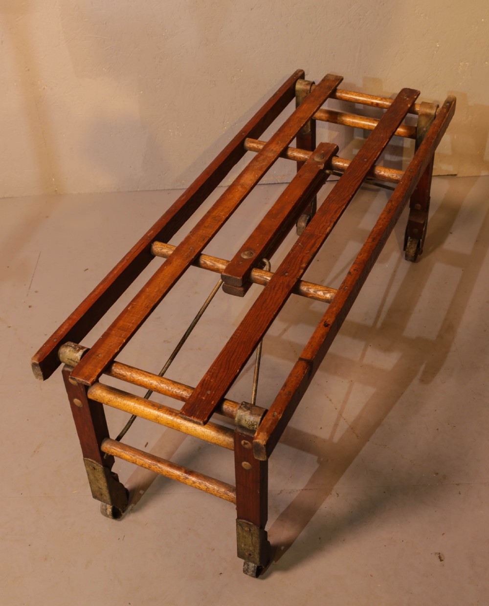 A slatted warm brown wooden luggage stand featuring metal castors and stamped maker details along the front rail. View VIII