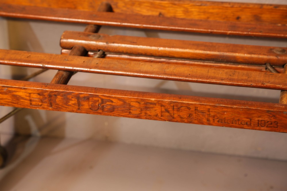 A slatted warm brown wooden luggage stand featuring metal castors and stamped maker details along the front rail. View IX