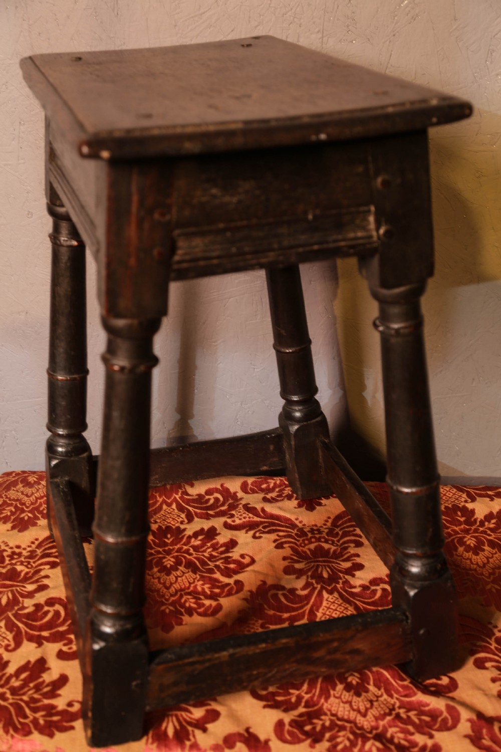 A seventeenth-century dark oak joint stool featuring turned legs, lower perimeter stretchers, and a flat top secured with wooden pegs.